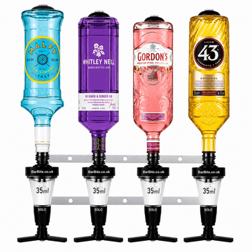Four liquor bottle dispensers mounted on a metal rail, each holding a different colored bottle: a blue Halcy gin bottle, a purple Whitley Nell gin bottle, a pink Gordon's gin bottle, and a gold 43 vodka bottle. The dispensers are branded BarBits.