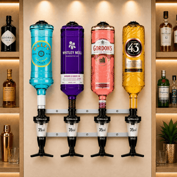 A wall-mounted bar display holding four liquor bottles on a metal rack: a turquoise bottle labeled Camp Cane, a purple bottle labeled Whitley Neill, a pink bottle labeled Gordon's, and a yellow bottle labeled 43. The bottles are mounted against a beige wall with built-in shelving behind them, and other bottles and glasses are visible on the shelves.