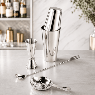 A polished silver cocktail bar set on a light marble countertop, including a shaker tin with lid, jigger, long bar spoon, strainer, and small pebble-surface spoon, with a softly blurred kitchen/bar background.
