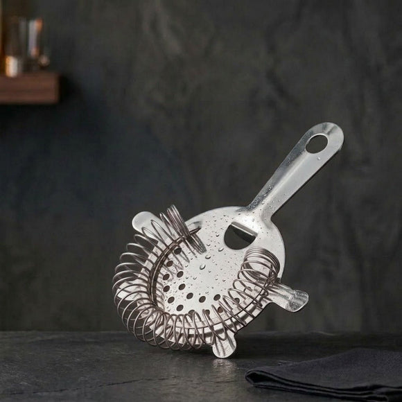 A stainless steel cocktail strainer with a wire mesh basket and angled handle, resting on a dark textured surface against a dark gray background.