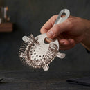 A hand holding a stainless steel cocktail strainer with a wire mesh filter and a hanging loop, set against a dark, moody background.