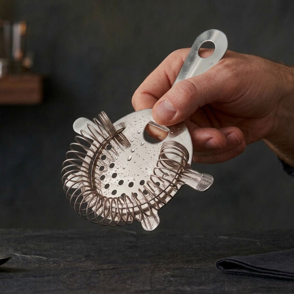 A hand holding a stainless steel cocktail strainer with a wire mesh filter and a hanging loop, set against a dark, moody background.