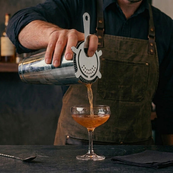 A bartender in a dark apron pours an amber cocktail from a metal shaker into a coupe glass, with a strainer on the shaker and bar tools on a dark countertop.