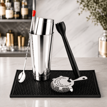 A stainless steel cocktail shaker set with a black rubber mat on a marble countertop. The set includes a metal shaker tin, a black strainer, a black muddler, a metal spoon with a red handle, and a metal bar spoon.