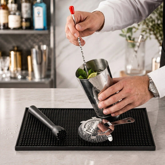 A person mixing a cocktail in a stainless steel shaker on a black bar mat, with a muddler and strainer nearby in a bright bar setting.