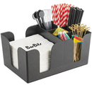 A black textured plastic bar organizer filled with white napkins, black straws, red-and-white striped straws, gold cocktail picks, and colorful mini umbrellas on a white background.