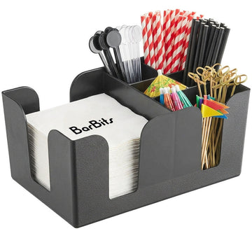 A black textured plastic bar organizer filled with white napkins, black straws, red-and-white striped straws, gold cocktail picks, and colorful mini umbrellas on a white background.