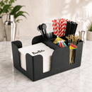 A black bar caddy holder organizer on a marble countertop, holding white napkins labeled BarBits, red-and-white striped straws, black straws, cocktail spoons, colorful picks, and gold-tone cocktail picks.