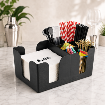 A black bar caddy holder organizer on a marble countertop, holding white napkins labeled BarBits, red-and-white striped straws, black straws, cocktail spoons, colorful picks, and gold-tone cocktail picks.
