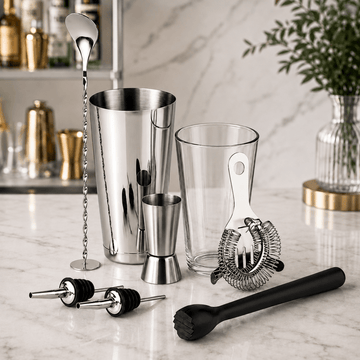 A cocktail bar tool set arranged on a white marble countertop, including a stainless steel shaker, mixing glass, jigger, strainer, muddler, and bar spoon, with a blurred kitchen background.