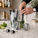 A set of stainless steel and glass cocktail bar tools on a marble countertop, including a metal shaker, jigger, strainer, muddler, and pourers, with a person's hands mudding lime into a glass.