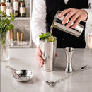 A bar tool set arranged on a marble countertop, including a stainless steel cocktail shaker, jigger, strainer, muddler, spoon, and muddler attachments. A person's hands are shown using the black muddler inside a glass with lime and mint.
