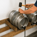 A person's hands use a rubber mallet to tap a stainless steel kegerator on a wooden rack against a white wall.