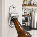 A silver metal bottle opener mounted on a white wall, with a brown glass beer bottle being opened. The background shows a blurred bar interior with bottles and stainless steel bar tools.