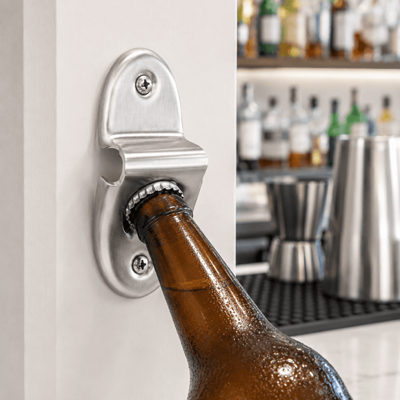 A silver metal bottle opener mounted on a white wall, with a brown glass beer bottle being opened. The background shows a blurred bar interior with bottles and stainless steel bar tools.