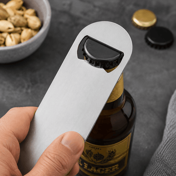 A hand holding a silver metal bottle opener with a black lever, positioned over an amber beer bottle with a gold cap on a dark gray textured surface.