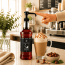Bottle of RealMint hazelnut syrup being poured into a latte with a hand pointing to it, set against a café background.
