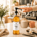 Vanilla syrup being poured into a cup of coffee with a vanilla bean and flower on a kitchen counter.