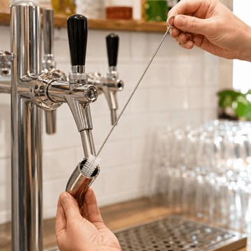 A close-up of a stainless steel beer tap cleaning brush being used to clean the inside of a metal tap handle, with a hand holding the handle and another hand pulling a metal cleaning rod. The background shows a blurred bar setting with glassware and a white subway tile wall.