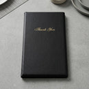 A black faux-leather bill presenter with 'Thank You' written in gold cursive on the front, resting on a gray textured surface with subtle dining props in the background.