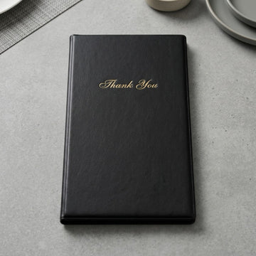 A black faux-leather bill presenter with 'Thank You' written in gold cursive on the front, resting on a gray textured surface with subtle dining props in the background.