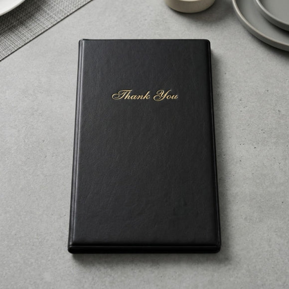 A black faux-leather bill presenter with 'Thank You' written in gold cursive on the front, resting on a gray textured surface with subtle dining props in the background.