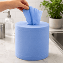 A blue paper towel dispenser on a white marble kitchen counter, with a hand pulling out a blue paper towel. A soap dispenser, a metal container, and a potted green plant are visible in the background.