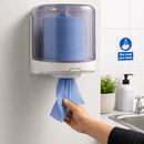 A wall-mounted dispenser holding a roll of blue tissue paper, with a hand pulling out a blue wipe. The scene is set in a clean bathroom with a sink, soap dispenser, and a hand-washing sign on the tiled wall.