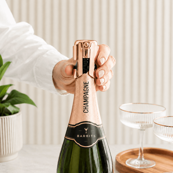 A hand in a white shirt is unscrewing the foil of a Barbits Champagne bottle. The green glass bottle has a metallic rose gold foil neck and a black label with gold lettering.