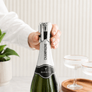 A hand with manicured nails holds a silver and black Barbitis Champagne bottle opener against the foil of a green champagne bottle. The scene is styled with a potted plant on the left and two coupe glasses on a wooden tray on the right, set against a clean, light beige ribbed wall background.