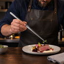 A close-up of a chef in a dark brown leather apron using clear glass tweezers to place a small green herb garnish on a plated dish of cooked meat, cream, and vegetables on a dark wooden table.