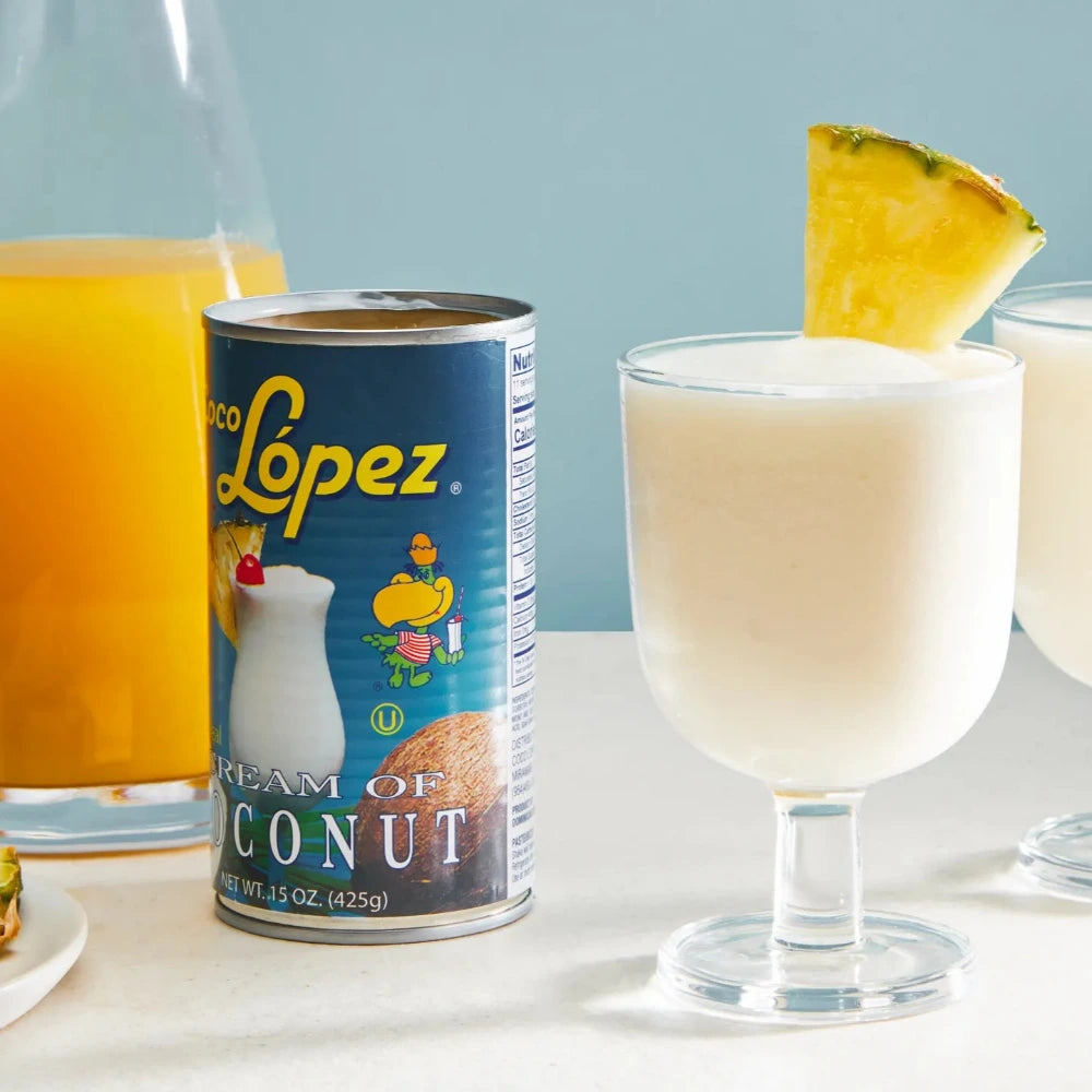 Two glasses of a creamy drink with pineapple slices, a can of Lopez brand coconut cream, and a bottle of orange juice on a light blue background.