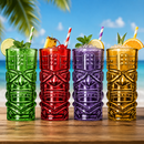 Four colorful tiki-style tumblers with a carved face design, filled with cocktails and garnished with citrus slices and mint, set on a wooden table with a beach and palm tree background.