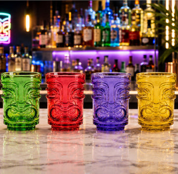 Four colourful tiki-style shot in green, red, purple, and yellow, arranged in a row on a polished marble bar counter with a blurred bar background.