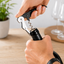A close-up of a person using a corkscrew to open a wine bottle, with a blurred indoor background showing a plant and wine glasses.