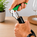 A close-up of a person using a green and silver wine bottle opener to remove a wine bottle cap on a wooden table, with a blurred plant and glass in the background.