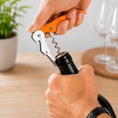 A close-up of a person's hands using an orange-handled corkscrew to open a black wine bottle on a wooden table, with blurred wine glasses and a potted plant in the background.
