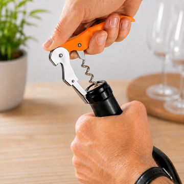 A close-up of a person's hands using an orange-handled corkscrew to open a black wine bottle on a wooden table, with blurred wine glasses and a potted plant in the background.