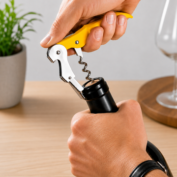 A close-up of a person using a yellow-handled corkscrew to open a black wine bottle on a wooden table, with a blurred plant and wine glass in the background.