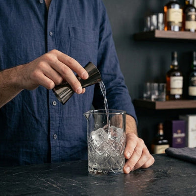 A person in a navy blue shirt pours a clear liquid from a black gunmetal jigger into a clear cut-glass mixing glass with ice, set on a dark bar counter with shelves of bottles in the background.