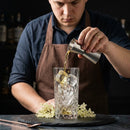 A bartender in a brown apron pours a clear amber liquid from a silver jigger into a faceted clear highball glass with ice, set on a dark table with small white flowers.