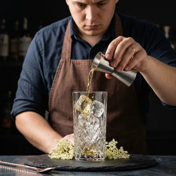 A bartender in a brown apron pours a clear amber liquid from a silver jigger into a faceted clear highball glass with ice, set on a dark table with small white flowers.