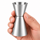 A stainless steel jigger with a brushed finish, held in a person's hand against a plain white background.