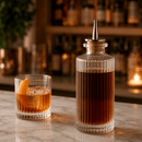 Bar setting with a glass of cocktail and a bottle of dark liquid on a marble surface.