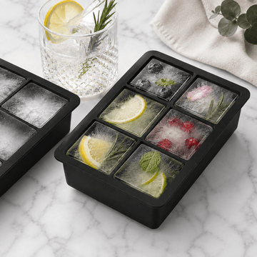 Black silicone ice cube tray with six compartments, shown filled with ice cubes containing lemon slices, mint, rosemary, blueberries, and cranberries. A matching tray is partially visible on the left, and a glass of iced drink with a lemon slice sits behind on a white marble surface with a cream cloth and greenery.