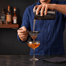 A person in a blue shirt pours an amber drink through a fine mesh gummetal strainer into a coupe glass with orange slices, with bar bottles blurred in the background.