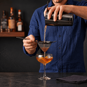 A person in a blue shirt pours an amber drink through a fine mesh gummetal strainer into a coupe glass with orange slices, with bar bottles blurred in the background.