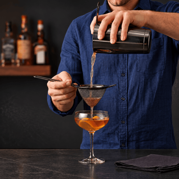 A person in a blue shirt pours an amber drink through a fine mesh gummetal strainer into a coupe glass with orange slices, with bar bottles blurred in the background.