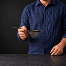 A man wearing a dark blue button-down shirt holds a small black mesh strainer with a metal handle over a dark surface against a dark background.