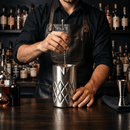 A bartender in a black shirt and dark brown leather apron stirs a cocktail in a textured stainless steel mixing tin on a dark bar counter, with bottles of liquor blurred in the background.